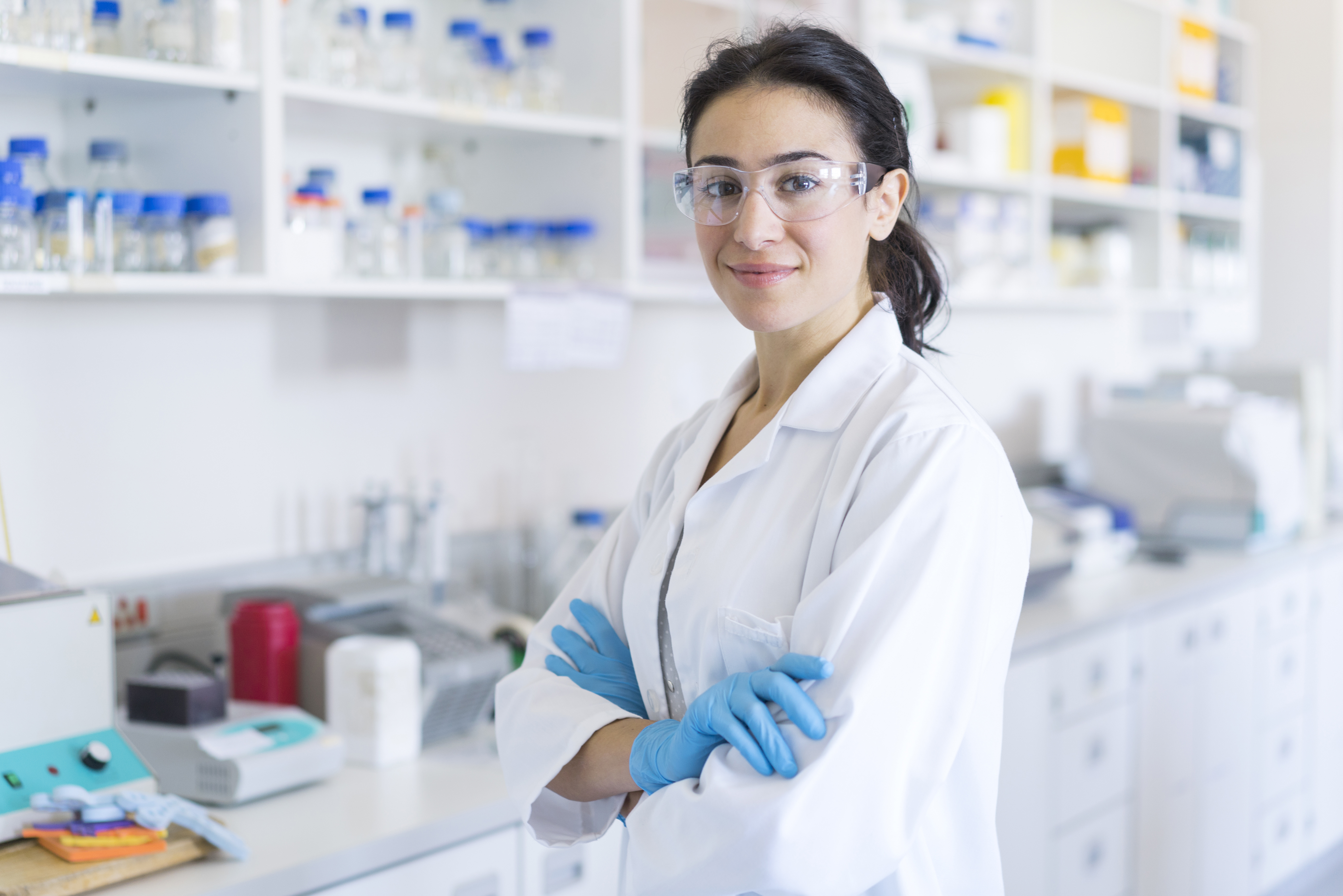 A photo of smiling female chemist by shelves. Portrait of confident scientist standing with arms crossed. She is wearing protective eyeglasses and lab coat at laboratory.